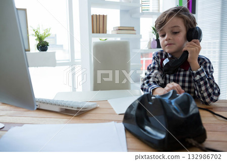 Boy sitting at desk in home office picking up telephone handset near computer monitor, copy space Boy sitting at desk in home office picking up telephone handset near computer monitor, copy space 132986702