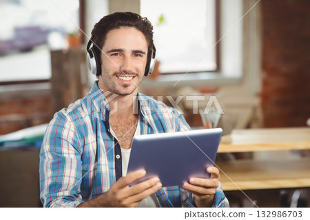 Man in twenties wearing headphones smiling toward camera while holding tablet at coworking office Man in twenties wearing headphones smiling toward camera while holding tablet at coworking office 132986703