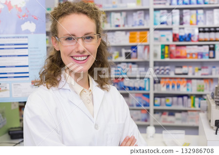Smiling female pharmacist standing behind counter in lab coat by stocked shelves, register, poster Smiling female pharmacist standing behind counter in lab coat by stocked shelves, register, poster 132986708