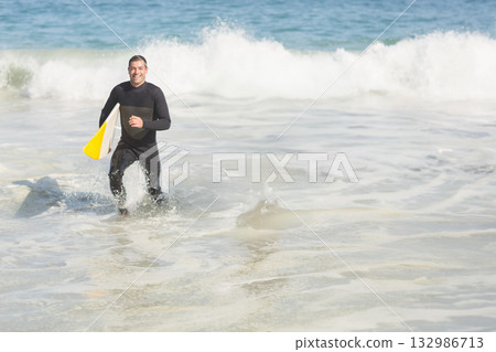 Middle aged man in black wetsuit emerging from ocean at beach carrying white and yellow surfboard 132986713