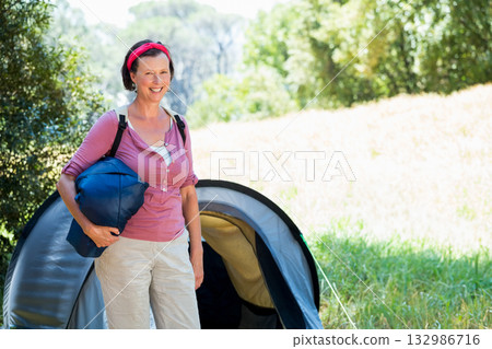 Senior woman in hiking gear carrying backpack and sleeping bag at forest campsite by pitched tent Senior woman in hiking gear carrying backpack and sleeping bag at forest campsite by pitched tent 132986716