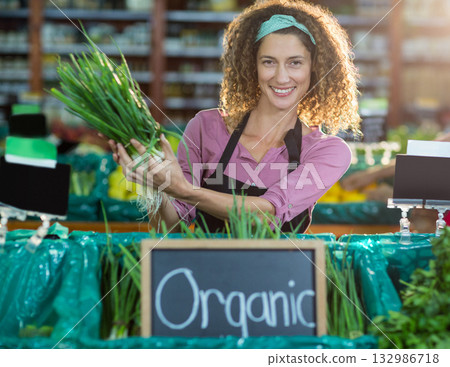 Female produce clerk wearing black apron smiling and holding green onions at market produce section 132986718