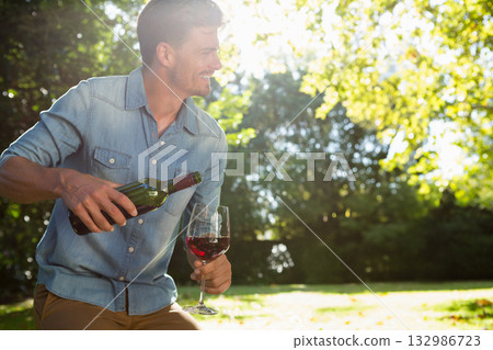 Man in denim shirt pouring red wine into glass on lawn under leafy trees, copy space Man in denim shirt pouring red wine into glass on lawn under leafy trees, copy space 132986723