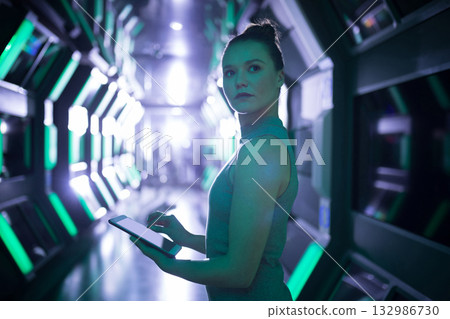 Woman in fitted grey dress holding tablet and scanning illuminated panels in spacecraft corridor 132986730