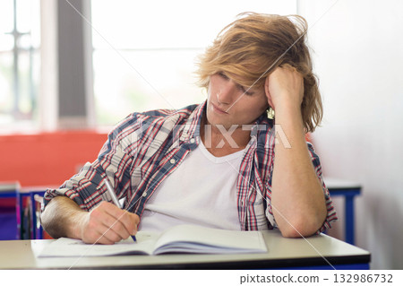 Male student writing in notebook with pen at desk in classroom under bright window light 132986732