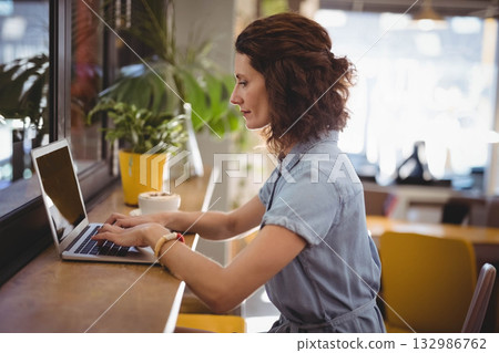 Woman working on laptop at wooden counter by window with yellow pot, white planter and wristwatch Woman working on laptop at wooden counter by window with yellow pot, white planter and wristwatch 132986762
