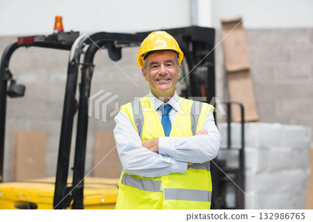 Man standing in warehouse wearing safety vest and hard hat by forklift with stacked pallets Man standing in warehouse wearing safety vest and hard hat by forklift with stacked pallets 132986765