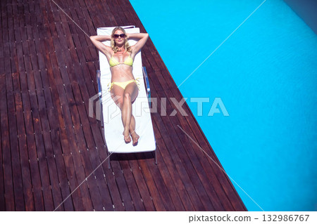 Woman reclining wearing yellow bikini and sunglasses on white lounge chair on deck beside blue pool 132986767