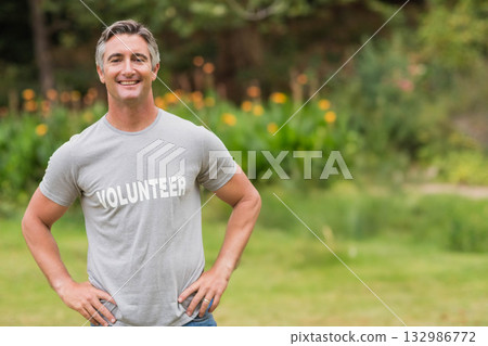 Middle-aged man standing with hands on hips in garden by flower bed wearing gray volunteer T-shirt 132986772