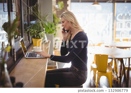 Woman juggling smartphone call and typing on laptop at cafe counter near coffee cup, copy space Woman juggling smartphone call and typing on laptop at cafe counter near coffee cup, copy space 132986779