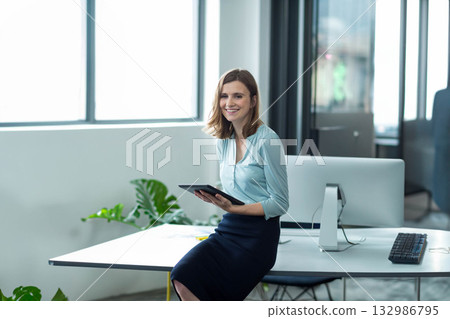 Woman sitting on edge of white desk holding tablet beside monitor, keyboard, potted plant in office Woman sitting on edge of white desk holding tablet beside monitor, keyboard, potted plant in office 132986795