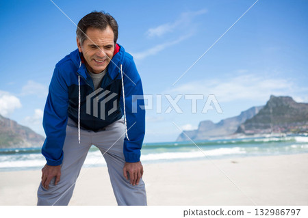 Senior man catching breath with hands on knees on beach wearing blue windbreaker and grey pants 132986797