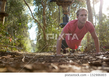 Man in red tee and shorts performing push-up on forest trail with wooden obstacle course 132986803