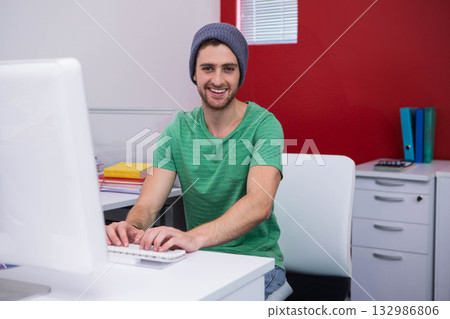 Man in twenties typing on desktop computer at modern office desk with notebooks and calculator Man in twenties typing on desktop computer at modern office desk with notebooks and calculator 132986806