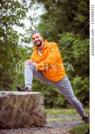 Man wearing bright orange windbreaker and sneakers stretching leg on boulder in forest clearing 132986811