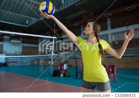 Female athlete standing near net holding volleyball overhead at gym beside red chairs, copy space Female athlete standing near net holding volleyball overhead at gym beside red chairs, copy space 132986815