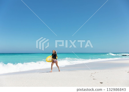 Female surfer wearing rash guard carrying yellow surfboard walking along beach, copy space 132986843