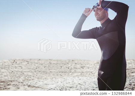 Male swimmer adjusting goggles on sandy beach in black wetsuit, blue swim cap, copy space Male swimmer adjusting goggles on sandy beach in black wetsuit, blue swim cap, copy space 132986844