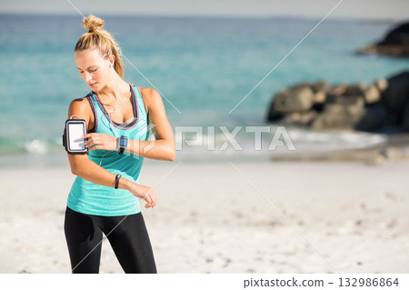 Woman in sportswear standing on shore by turquoise sea, adjusting smartphone armband, copy space 132986864