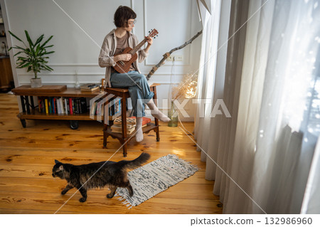 Focused young woman rehearsing ukulele by window, immersed in chord repetition with cat walk around Focused young woman rehearsing ukulele by window, immersed in chord repetition with cat walk around 132986960