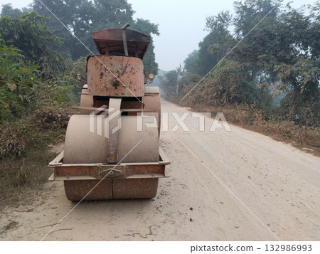 Rear view of an old rusty road roller parked on a dusty rural road during construction, showing double metal wheels used for compacting surface layers in village infrastructure work 132986993