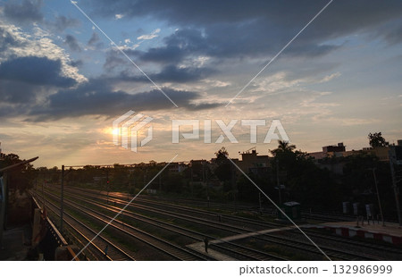 Cloudy evening sky with sun setting over railway lines and silhouetted buildings in the distance 132986999