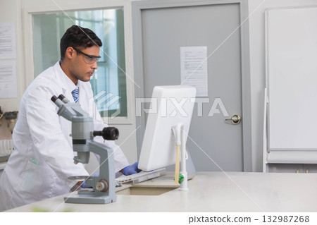 Male researcher wearing lab coat, gloves typing on PC at lab bench with microscope, pipette stand 132987268