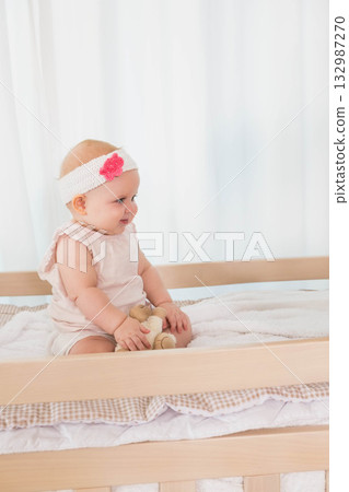 Child baby girl sitting on changing table in nursery wearing pink onesie holding plush teddy bear Child baby girl sitting on changing table in nursery wearing pink onesie holding plush teddy bear 132987270