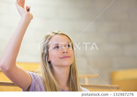 Teenage female student raising hand and looking upward in lecture hall with ballpoint pen 132987276