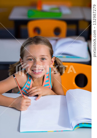 Smiling school girl writing in notebook at classroom desk with pencil and orange chairs Smiling school girl writing in notebook at classroom desk with pencil and orange chairs 132987279