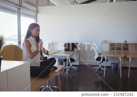 Asian woman meditating cross-legged on wooden desk beside pink telephone in office, copy space 132987282