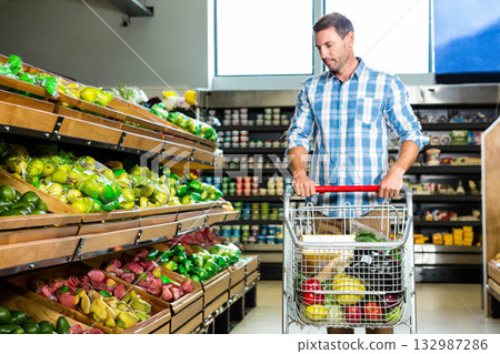 Mid adult man pushing shopping cart of produce by wooden bins in supermarket aisle, copy space Mid adult man pushing shopping cart of produce by wooden bins in supermarket aisle, copy space 132987286