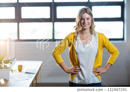 Woman standing with hands on hips in modern office at desk with computer, plant and mug Woman standing with hands on hips in modern office at desk with computer, plant and mug 132987291