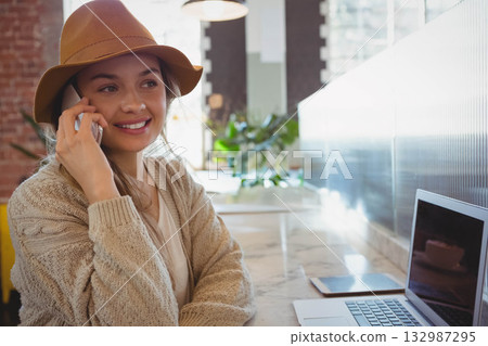 Woman wearing fedora calling on smartphone near laptop and tablet at cafe counter, copy space Woman wearing fedora calling on smartphone near laptop and tablet at cafe counter, copy space 132987295