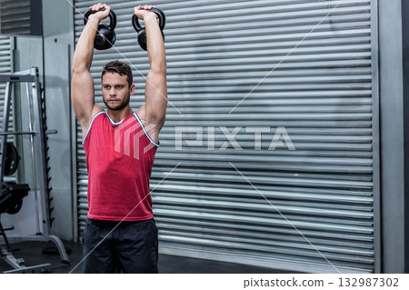 Male athlete in red shirt lifting kettlebells at gym with weight rack and metal shutter door Male athlete in red shirt lifting kettlebells at gym with weight rack and metal shutter door 132987302