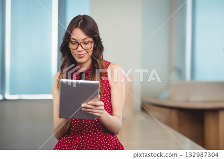 Woman wearing eyeglasses holding tablet and reading screen in office conference area with blinds 132987304