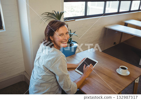 Senior woman sitting at wooden desk in bright office using tablet near potted plant, copy space 132987317