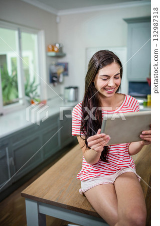 Woman sitting on wooden kitchen island holding tablet in home kitchen near potted plant, copy space 132987318