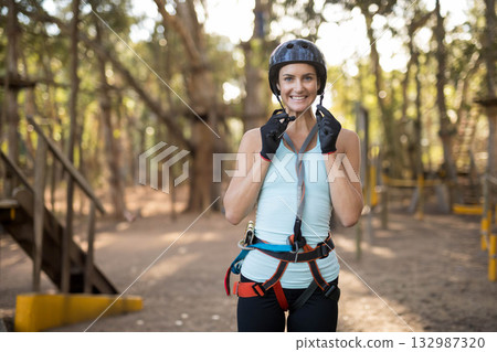Woman adjusting chin strap wearing protective gloves on forest ropes course with helmet and harness 132987320