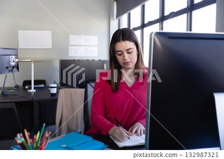 Woman office worker wearing magenta blouse writing in notebook at desk with takeaway coffee cup 132987321
