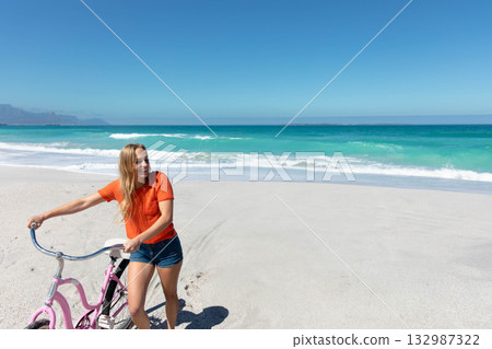 Standing woman wearing orange T-shirt and shorts beside pink cruiser bicycle on sandy beach 132987322