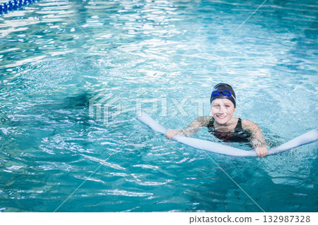 Woman floating in clear blue pool wearing black swimsuit swim cap gripping foam noodle, copy space Woman floating in clear blue pool wearing black swimsuit swim cap gripping foam noodle, copy space 132987328
