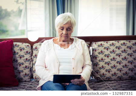Senior woman holding black tablet while sitting on floral sofa in living room near light curtains Senior woman holding black tablet while sitting on floral sofa in living room near light curtains 132987331