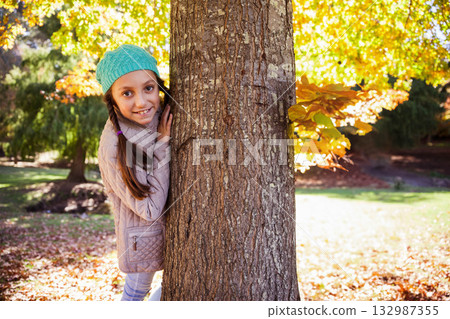 Female teenage girl peeking behind trunk in park wearing teal hat, beige quilted jacket, copy space Female teenage girl peeking behind trunk in park wearing teal hat, beige quilted jacket, copy space 132987355