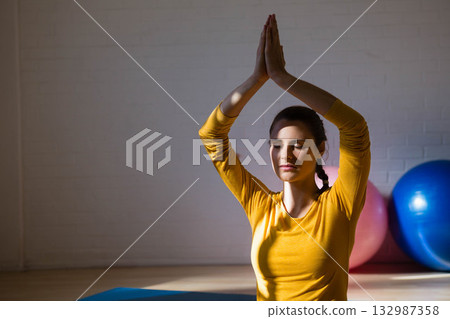 Female yoga practitioner sitting cross-legged on blue mat in studio with exercise balls, copy space Female yoga practitioner sitting cross-legged on blue mat in studio with exercise balls, copy space 132987358