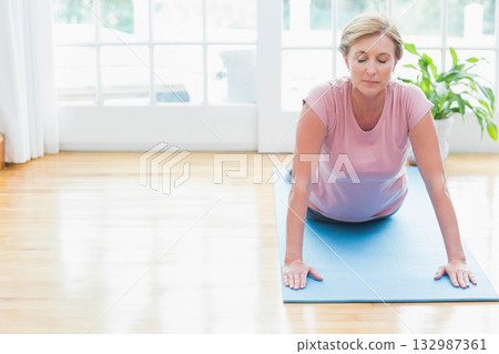 Woman stretching on blue mat in bright living room beside green potted plant, copy space Woman stretching on blue mat in bright living room beside green potted plant, copy space 132987361