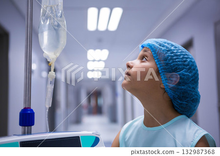 Female child wearing gown and cap standing in hospital corridor observing pump's IV bag, copy space 132987368