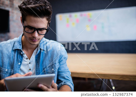 Man wearing eyeglasses sitting at office table holding tablet viewing sticky note whiteboard Man wearing eyeglasses sitting at office table holding tablet viewing sticky note whiteboard 132987374
