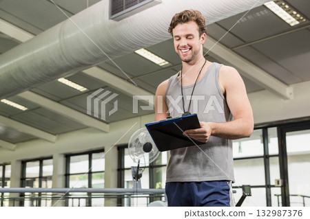 Male trainer in sportswear holding clipboard checking notes in gym with treadmills, weight benches 132987376