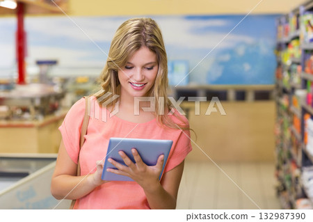 Woman in her twenties holding tablet and carrying bag scanning shelves in supermarket aisle 132987390